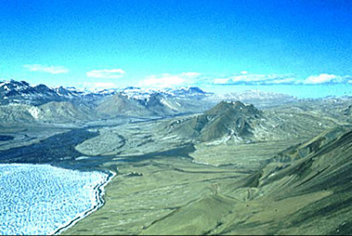 Figure 13: Axel Heiberg Island, Nunavut - This photograph shows a bay with a flat terrain surrounding the water. The water is covered with ice. There is no tree. A river is flowing into the bay. In the background, there are snowy mountain peaks.