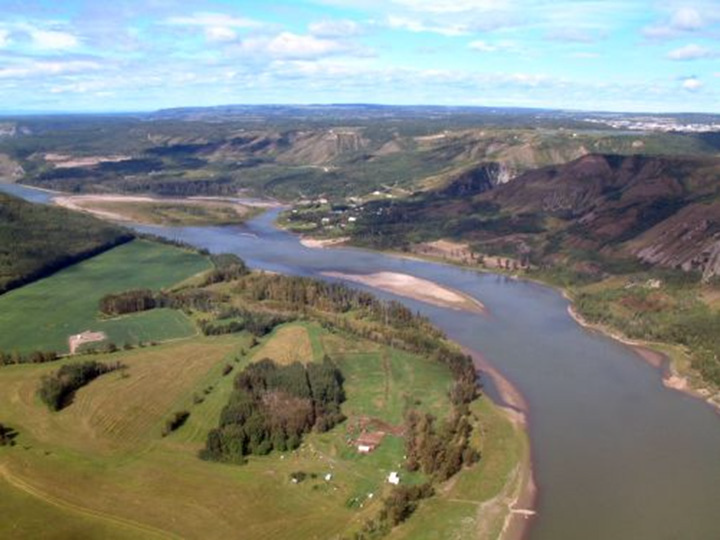 Figure 20: Peace River, British Columbia - This photograph shows a river flowing on a flat large valley surrounded by low plateaus. The valley and the plateaus are covered with vegetation made of grass, shrubs and trees. There is a populated place in the background, to the right, and homesteads can be seen along the river.