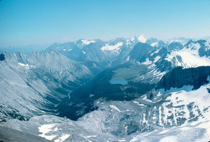 Figure 24: Upper Kananaskis River, Alberta - This photograph shows a deep U-shape valley surrounded by high knife-edge peaks and ridges. The bottom of the valley is covered by vegetation and a river goes through. The left slope is steep and cover with rock. The right slope is divided in two parts. The bottom part is steep and covered with vegetation, then there is a plateau with a small lake on it, and the top part of the slope is step and ends with snowed peaks. In the background there are several ridges of snowy peaks and a blue sky without clouds.